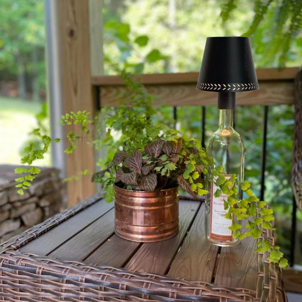 Flaschenaufsatzleuchte auf leerer Weinflasche neben Zimmerpflanze im Kupfertopf auf Rattan-Gartentisch