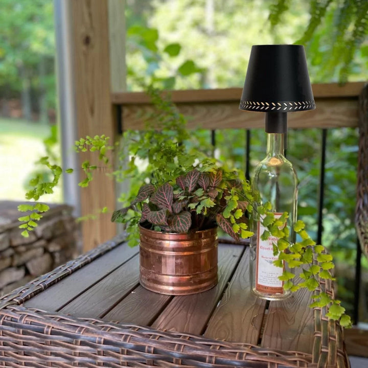 Flaschenaufsatzleuchte auf leerer Weinflasche neben Zimmerpflanze im Kupfertopf auf Rattan-Gartentisch