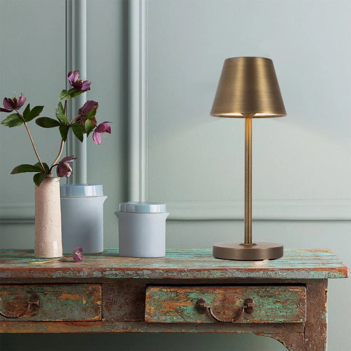 Kabellose LED-Akkuleuchte in Bronze auf antikem Vintage-Holztisch neben Blumenvase