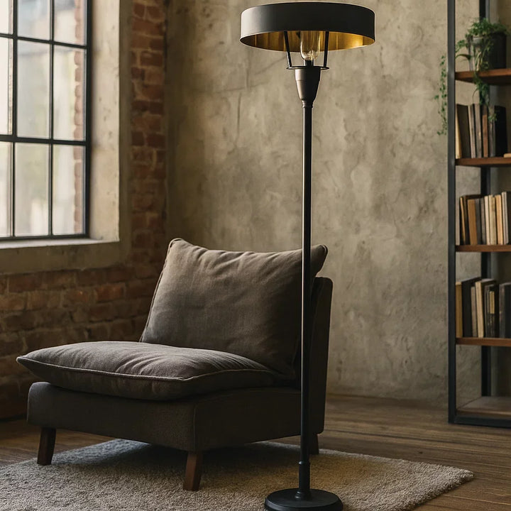 Eingeschaltete Art-Déco-Stehlampe in Mattschwarz mit goldglänzender Schirminnenseite neben grauem Lounge-Sessel in modernem Loft vor Beton- und Ziegelwand