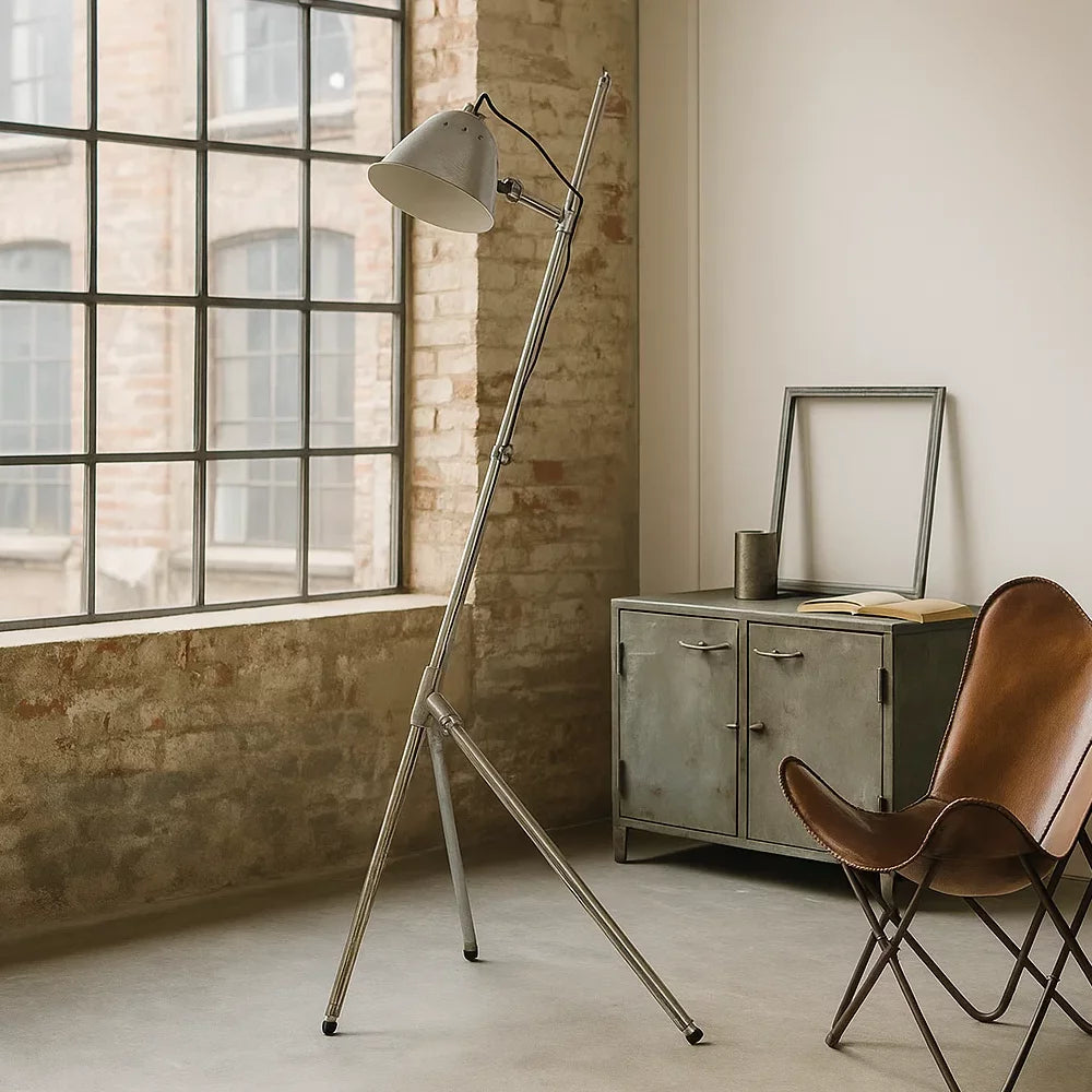 Verstellbare Stativ-Stehleuchte in Antiksilber vor Ziegelwand neben Metallsideboard und Butterfly-Ledersessel