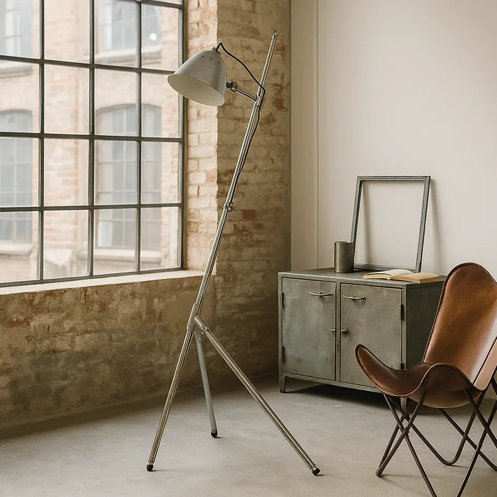 Verstellbare Stativ-Stehleuchte in Antiksilber vor Ziegelwand neben Metallsideboard und Butterfly-Ledersessel