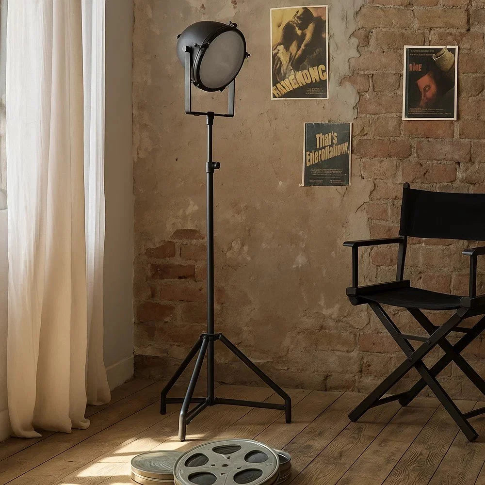 Eingeschaltete Dreibein-Stehlampe in Mattschwarz mit Scheinwerferkopf neben schwarzem Regiestuhl vor Backsteinwand im Filmset-Ambiente