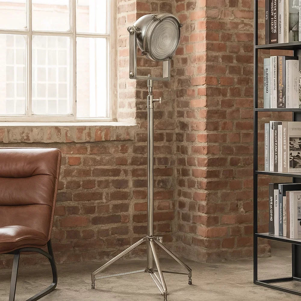 Eingeschaltete Dreibein-Stehlampe in Antiksilber vor Klinkerziegelwand neben braunem Ledersessel und schwarzem Bücherregal