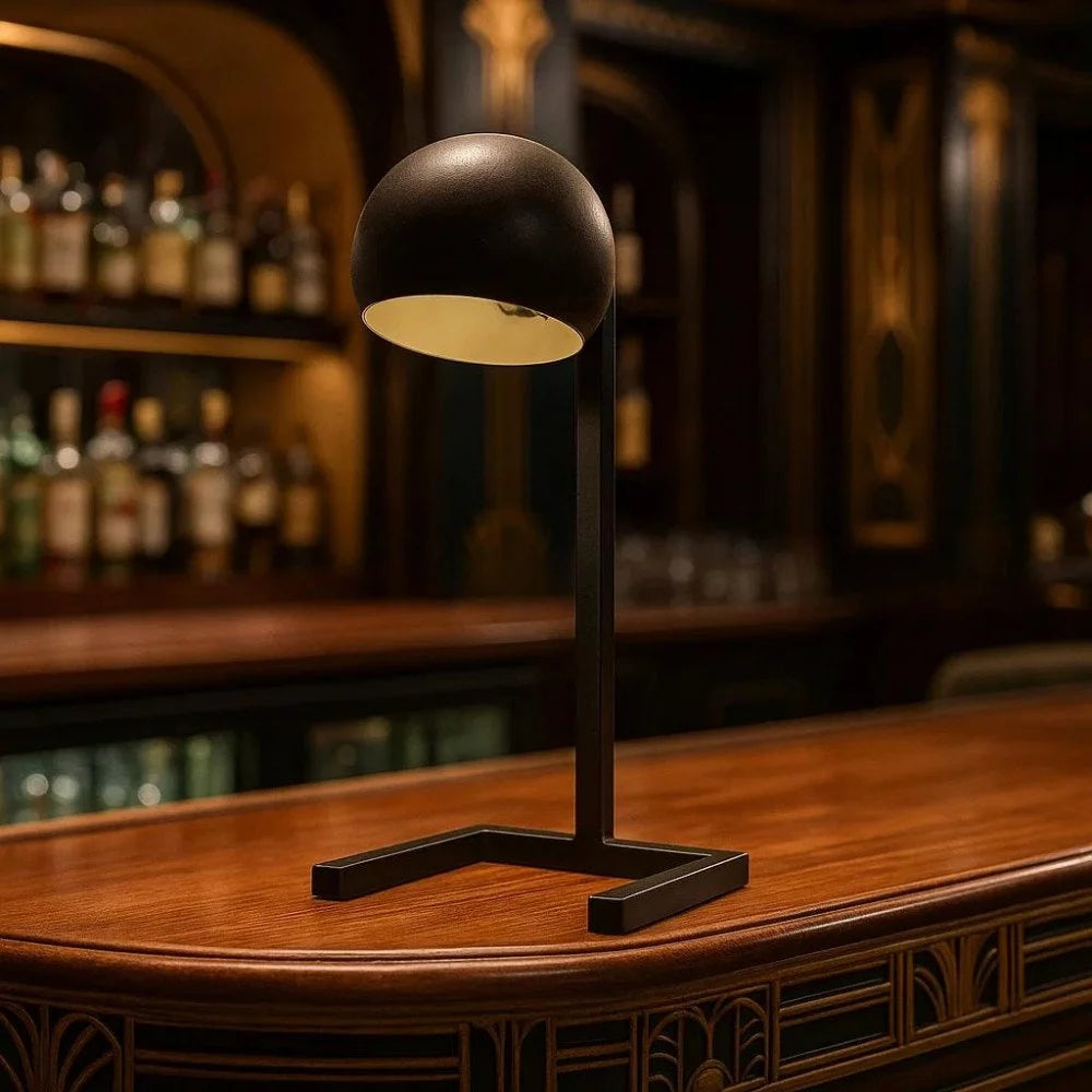 Mattschwarze Mid-Century-Tischleuchte mit neigbarem Kugelkopf eingeschaltet auf Holztheke einer Bar
