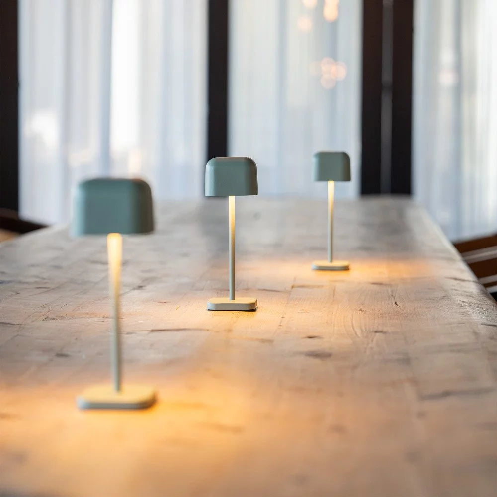 Drei kabellose Tischlampen Cabo Small Grün Grau hintereinander auf Holztisch – Stimmungslicht