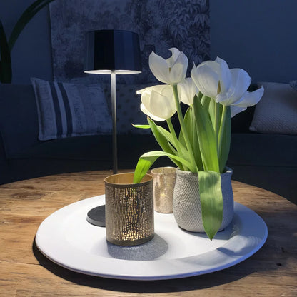 Kabellose Tischlampe Lido Anthrazit leuchtend auf weißem Teller mit Blumen – romantisches Stimmungslicht
