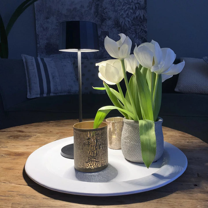 Kabellose Tischlampe Lido Anthrazit leuchtend auf weißem Teller mit Blumen – romantisches Stimmungslicht
