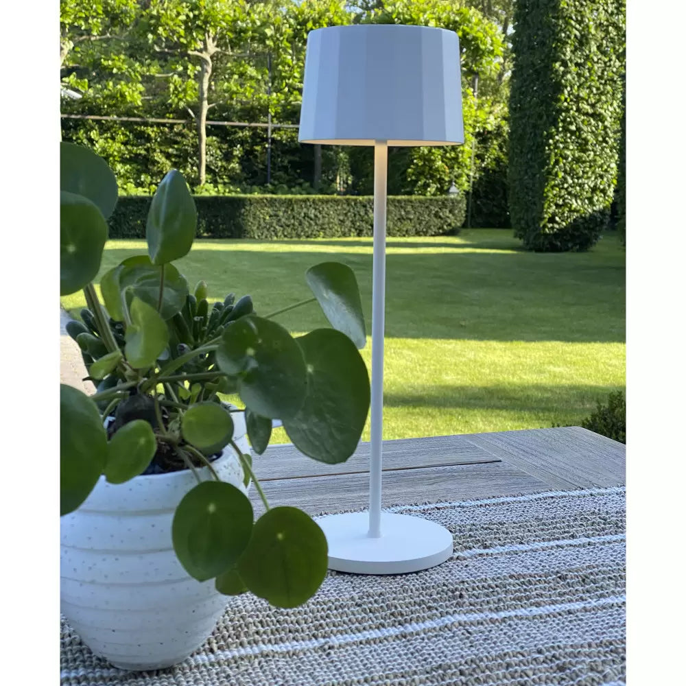 Moderne weiße Outdoor-Akkuleuchte auf Terrassentisch mit Tischläufer vor grünem Gartengrundstück