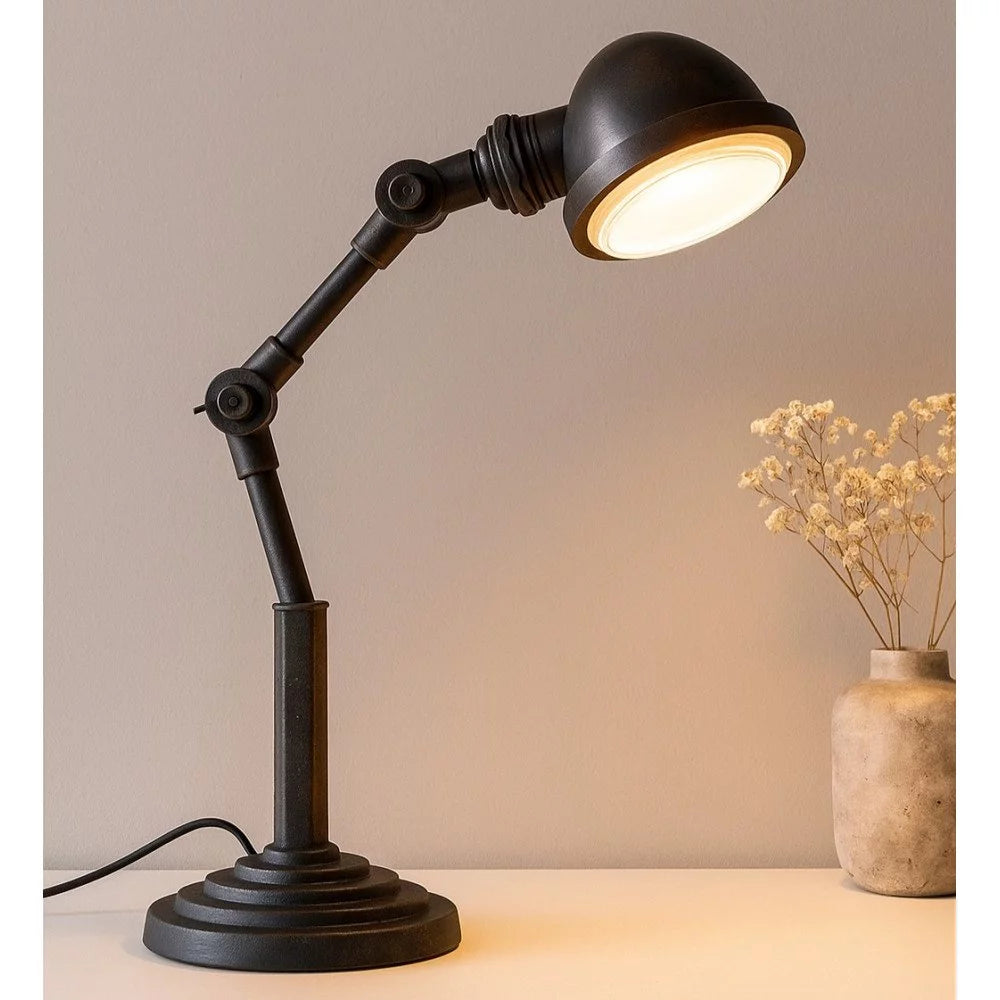 Mattschwarze Vintage-Schreibtischlampe mit leuchtender Glaslinse auf hellem Tisch neben Vase mit Trockenblumen