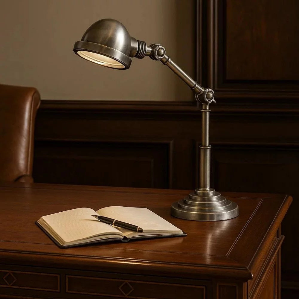 Klassische Gelenkarm-Schreibtischlampe in Antiksilber mit Glaslinse eingeschaltet über Notizbuch auf edlem Holzschreibtisch