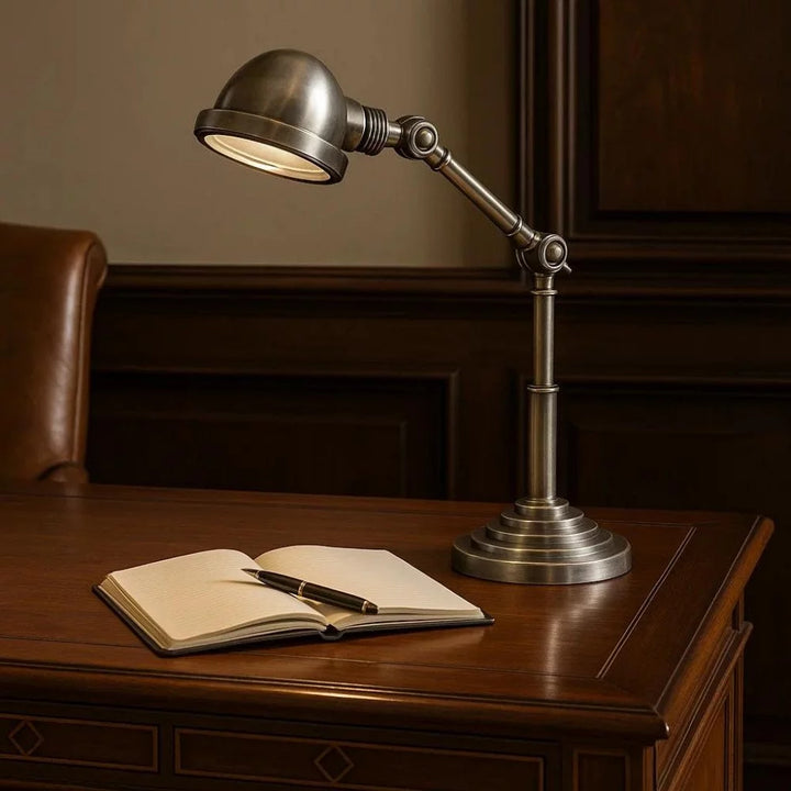 Klassische Gelenkarm-Schreibtischlampe in Antiksilber mit Glaslinse eingeschaltet über Notizbuch auf edlem Holzschreibtisch