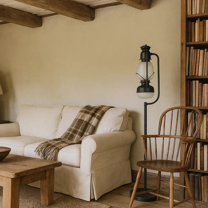 Schwarze Vintage-Laternen-Stehlampe mit Opalglaszylinder neben hellem Stoffsofa, Holzstuhl und Bücherregal im Landhaus-Wohnzimmer