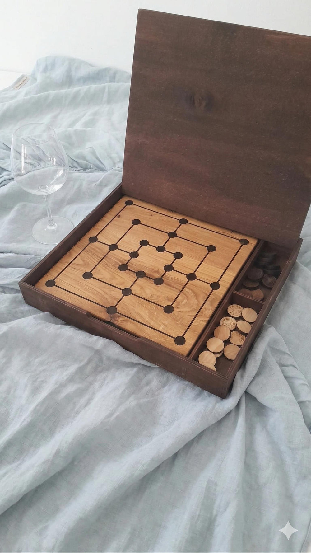 Geöffnete dunkel gebeizte Holzschachtel des Mühlespiels aus Eiche mit Spielbrett und Spielsteinen neben einem Weinglas