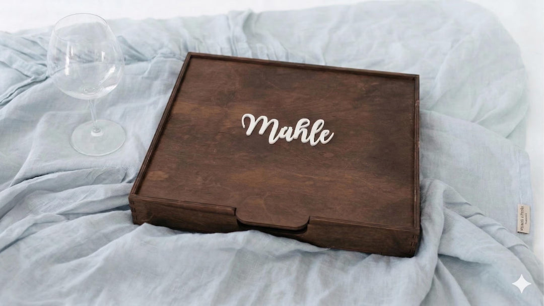 Geschlossene dunkle Holzschachtel mit Schriftzug „Mühle" auf hellblauem Leinentuch neben einem Weinglas