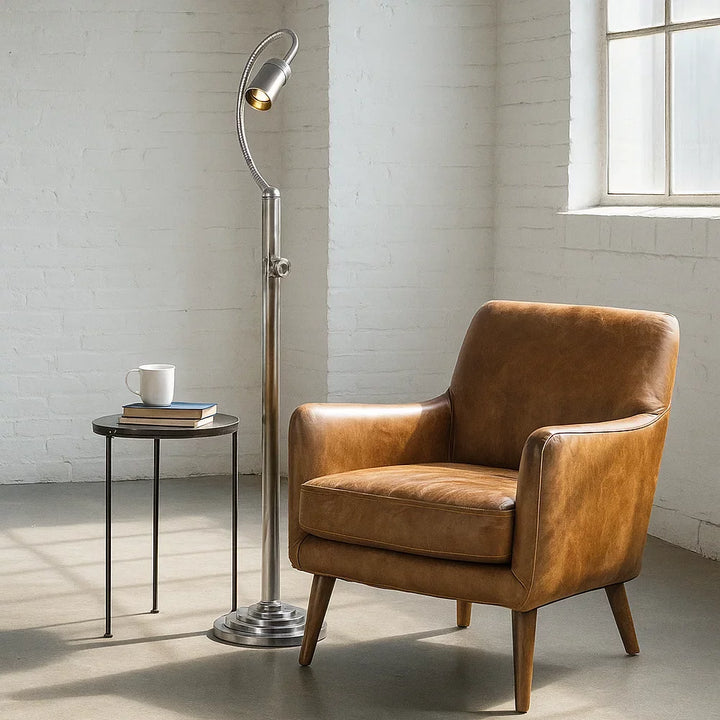 Eingeschaltete Vintage-Lese-Stehlampe in Antiksilber mit flexiblem Schwanenhals neben cognacbraunem Ledersessel im Loft-Ambiente