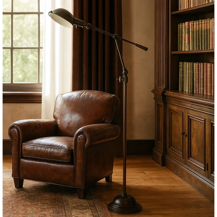 Eingeschaltete Stehleuchte mit Metallschirm in warmer Antikbronze neben braunem Chesterfield-Ohrensessel aus Leder vor dunklem Bibliotheksregal