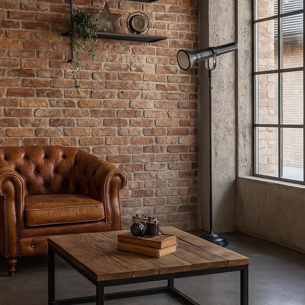 Industrielle Stehlampe in Mattschwarz im Design einer historischen Stabtaschenlampe neben braunem Chesterfield-Ledersessel vor roter Backsteinwand im Loft