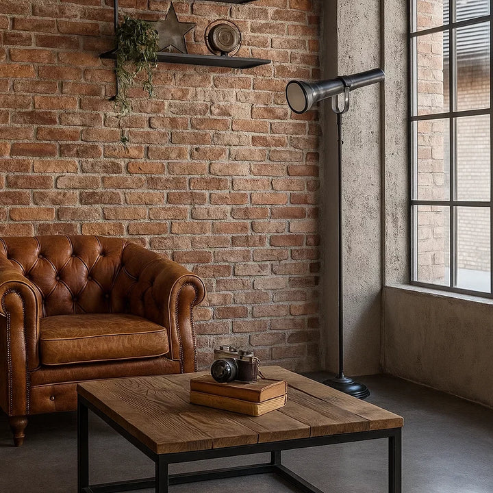 Industrielle Stehlampe in Mattschwarz im Design einer historischen Stabtaschenlampe neben braunem Chesterfield-Ledersessel vor roter Backsteinwand im Loft