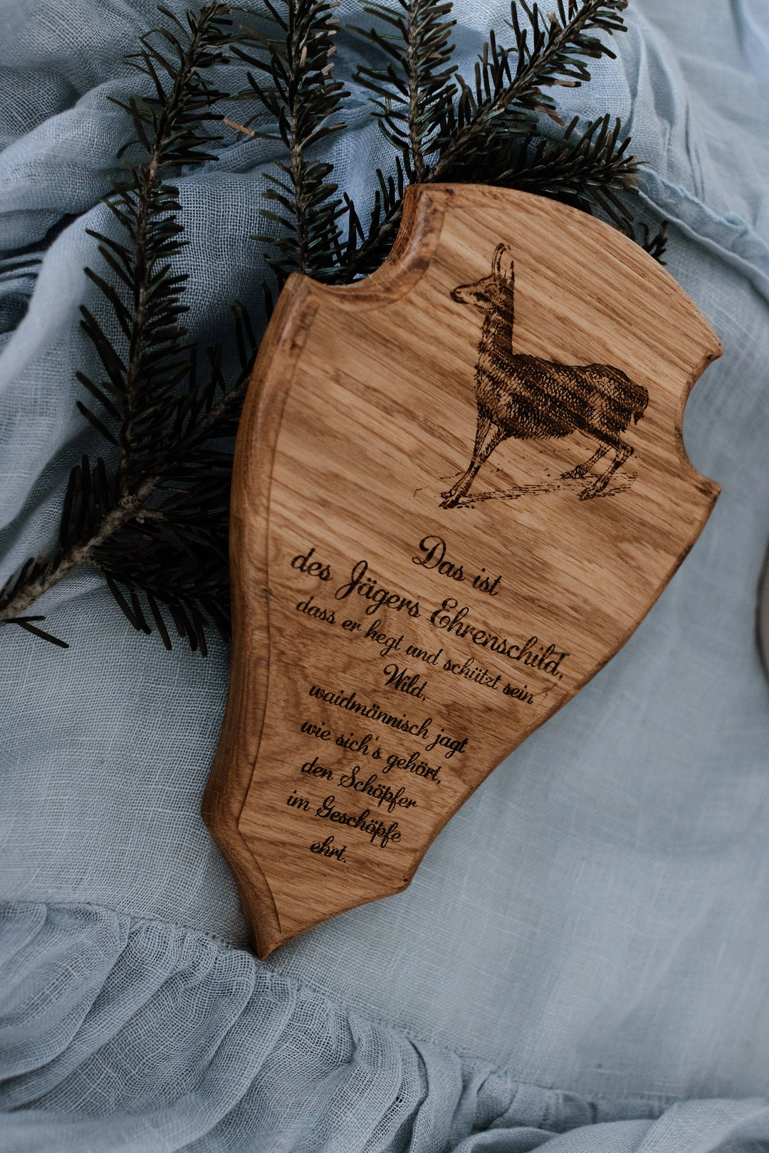 Trophäenschild aus massiver Eiche mit graviertem Gams-Motiv und Jägerspruch auf hellblauem Leinentuch